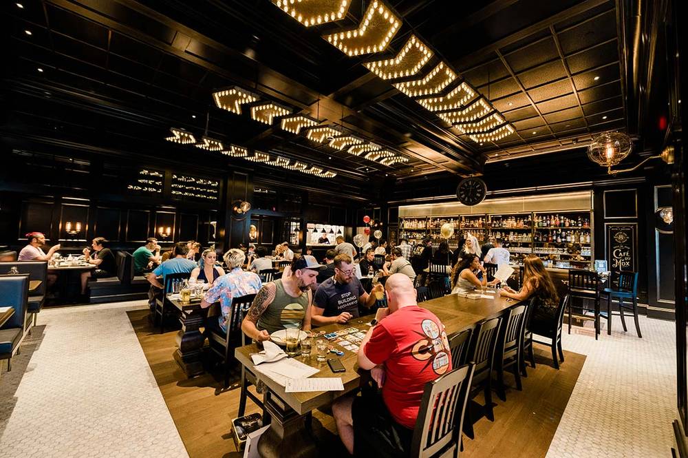 The dining room at Mox Boarding House, with groups playing board games at long wooden tables under rows of bulb lighting